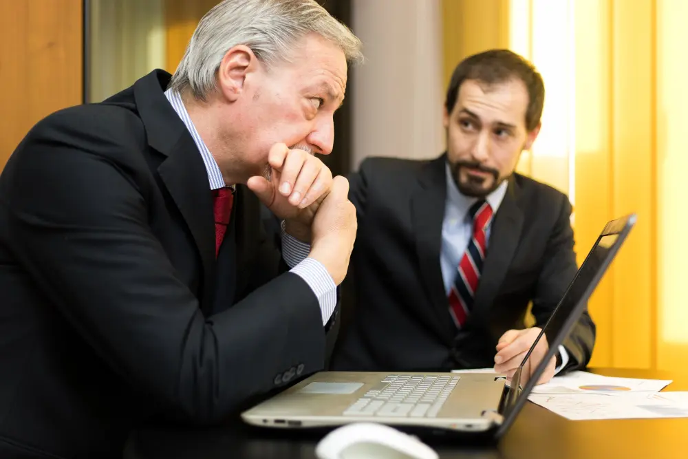 An older businessman listens intently with his hand to his chin while an advisor points to a laptop screen during a serious financial consultation, representing expert bankruptcy lawyers.