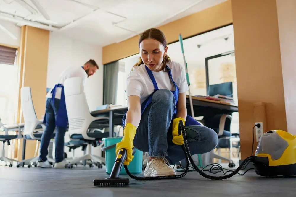 A professional cleaning company crew member in a blue apron and yellow gloves vacuums an office floor while a coworker cleans a desk workspace in the background.