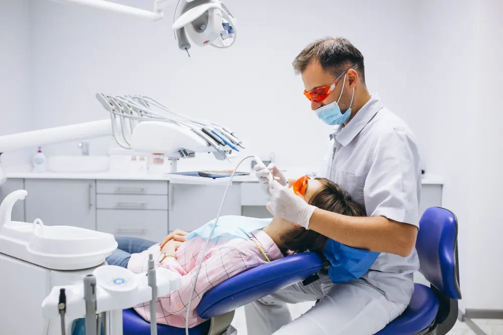 A male dentist wearing protective glasses, a mask, and gloves performs a dental procedure on a patient in a modern clinic chair, representing experienced professional dentists.
