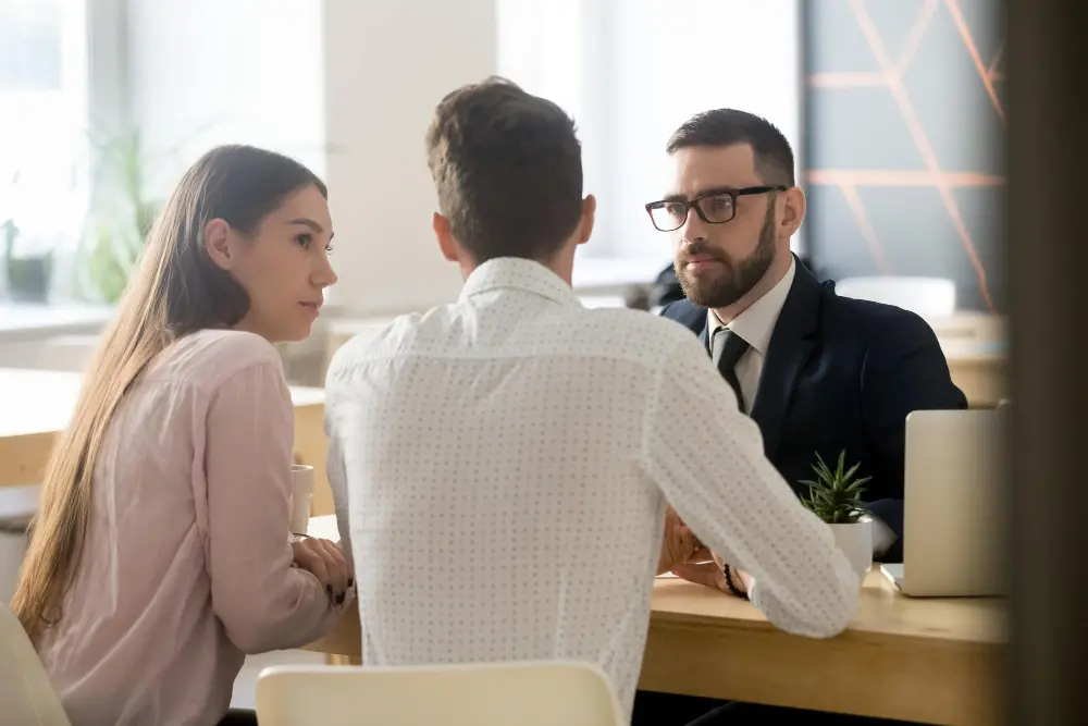 A male attorney with glasses and a suit listens attentively to a couple sitting across a wooden office table during a consultation, representing an experienced divorce lawyer.