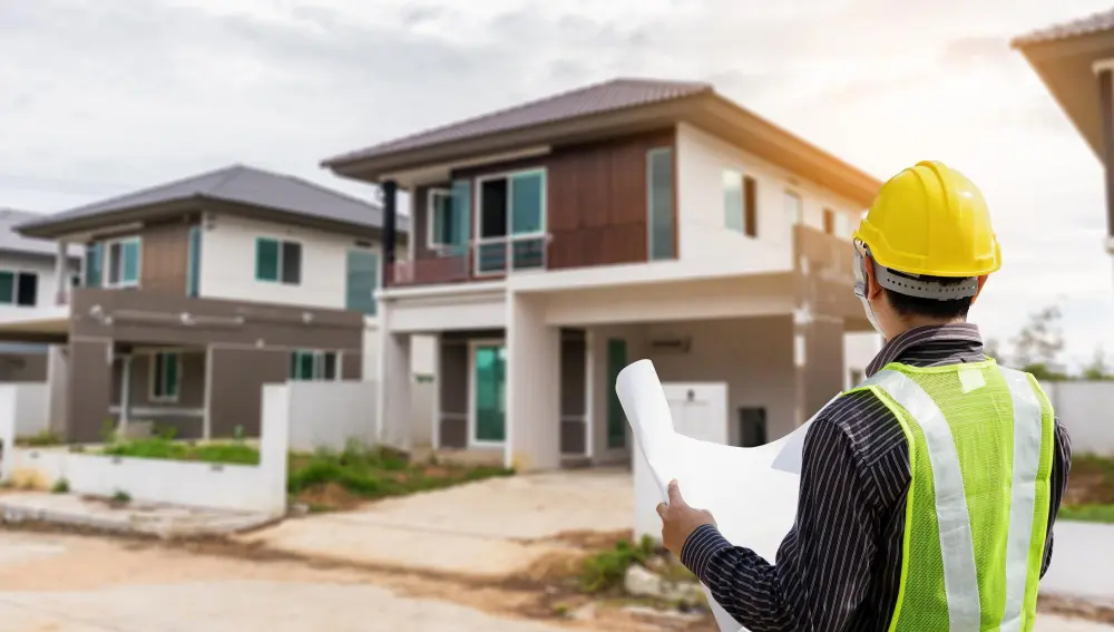 A construction engineer in a yellow hard hat and safety vest holds architectural blueprints while looking at a row of modern two-story houses, representing professional home builders.