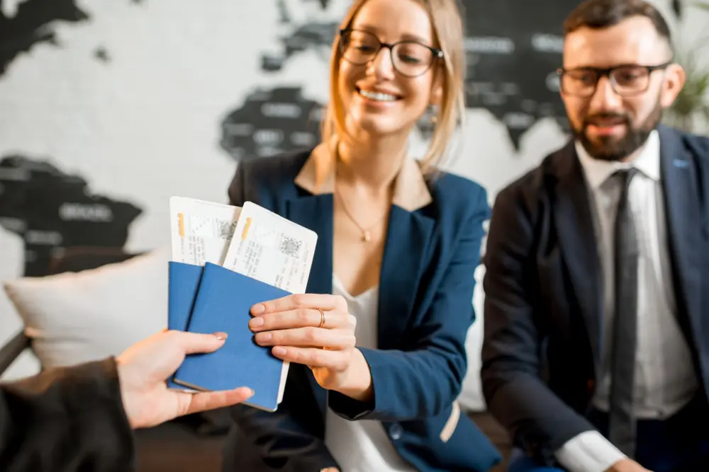 A smiling female attorney in a blue blazer hands over passports and boarding passes to a client next to a colleague in a suit, representing professional immigration lawyers.