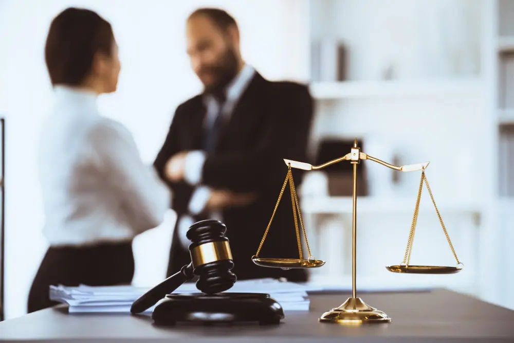 A golden scale of justice and a wooden judge gavel sit on an office desk with corporate legal professionals discussing a case out of focus, representing professional lawyers.