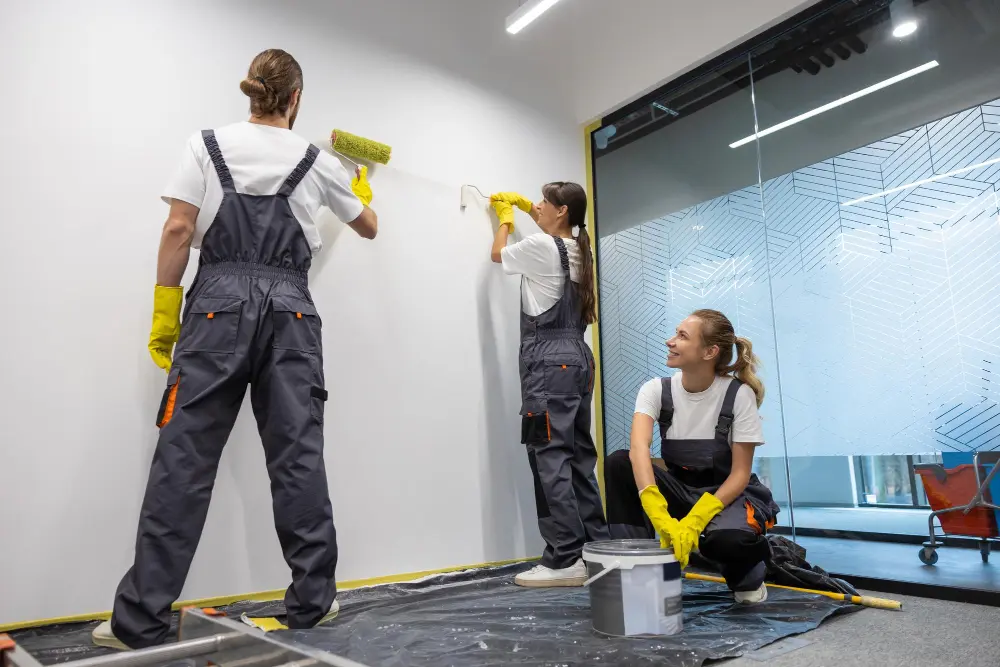 A professional crew in grey overalls and yellow gloves uses rollers to paint a white wall indoors near a paint bucket and drop cloth, representing top painting companies.