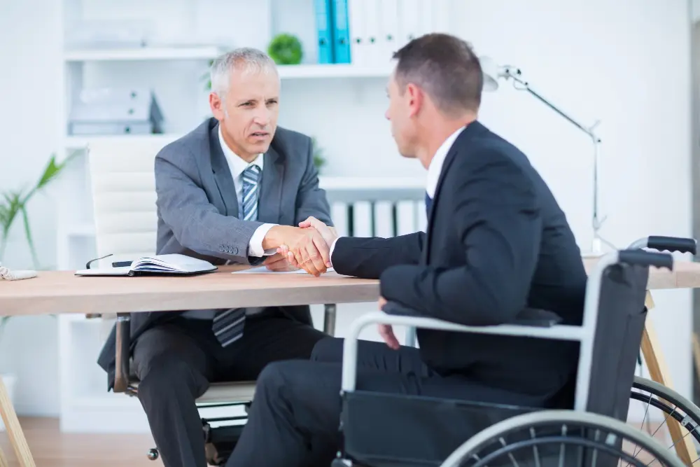 A male attorney in a grey suit shakes hands across a desk with a client sitting in a wheelchair during a legal consultation, representing dedicated personal injury lawyers.