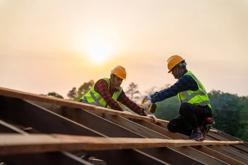 Two roofers wearing yellow hard hats and high-visibility safety vests use a power drill on a wooden residential frame at sunset, representing a professional roofing company.